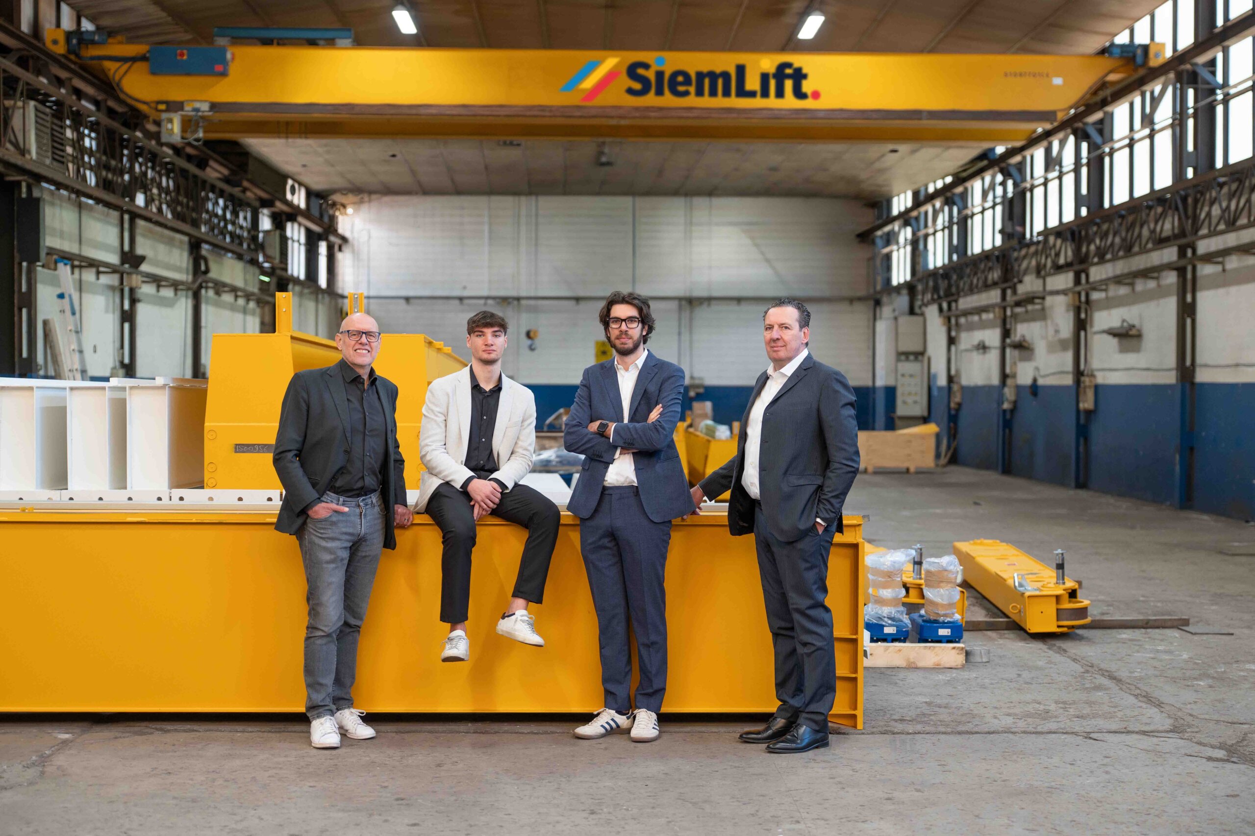 Quattro componenti del team SiemLift in posa all'interno dello stabilimento davanti a una gru a ponte gialla con marchio aziendale.